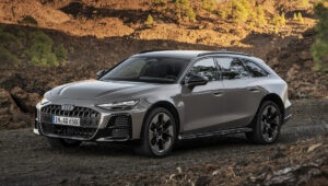 Audi A6 allroad cinza em primeiro plano, parada sobre terreno rochoso, vista dianteira e lateral.