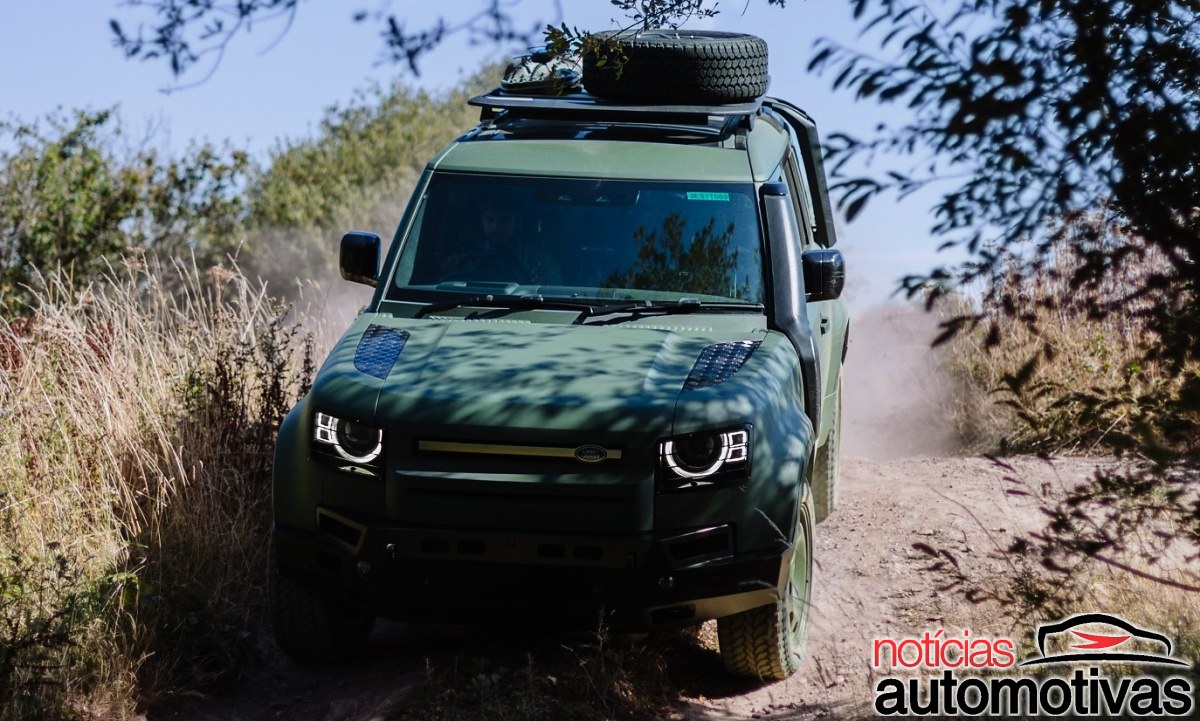 SUV verde-escuro da Defender visto de frente em estrada estreita, com estepe no teto e poeira ao fundo.