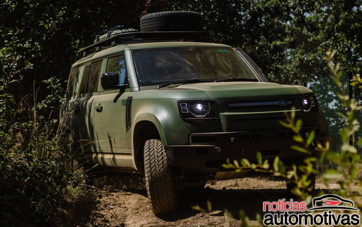 SUV verde-escuro da Defender em estrada de terra, visto de frente e pela lateral, com pneus fora de estrada e estepe no teto.