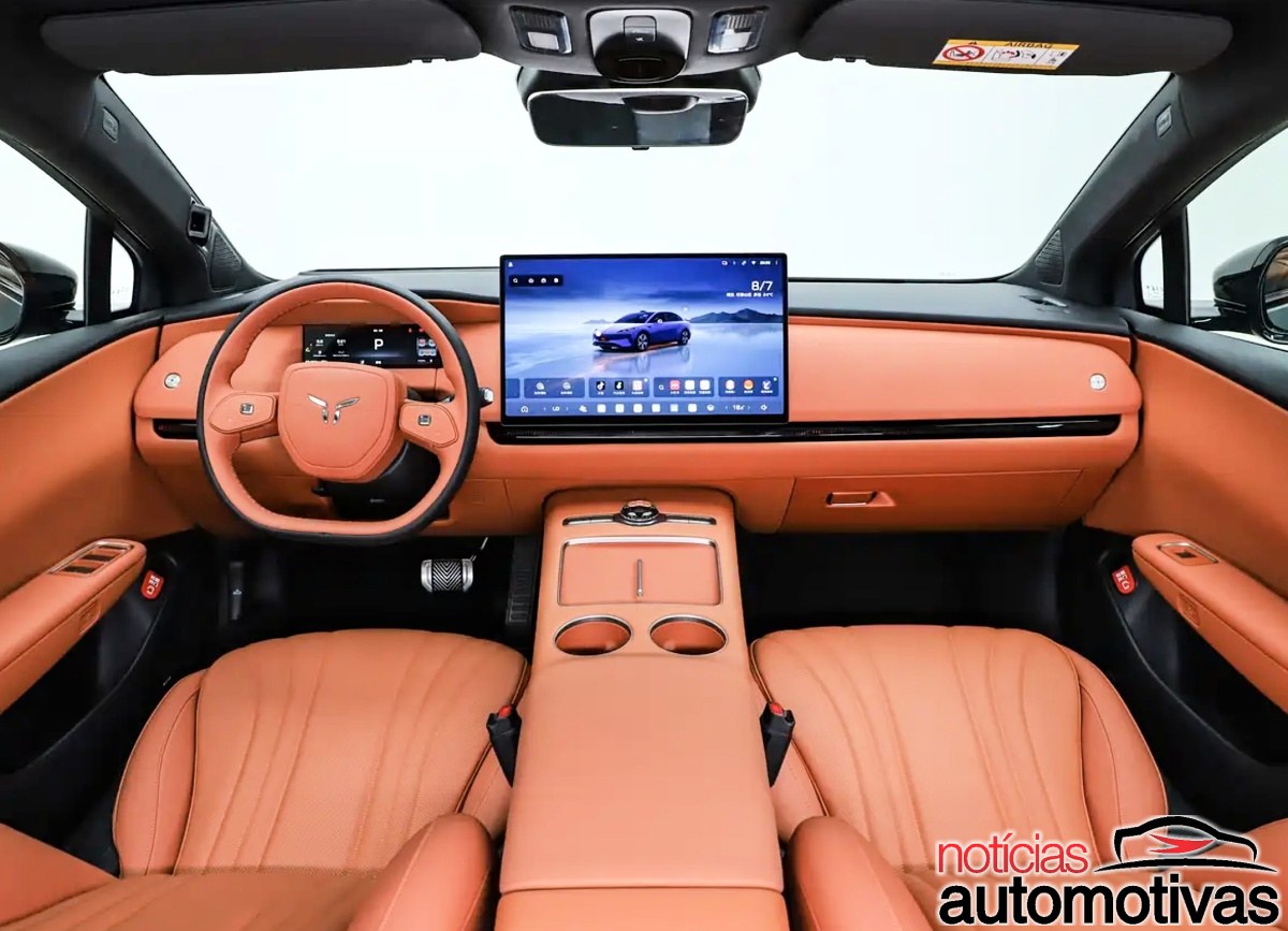 Interior dianteiro com bancos e painel revestidos em laranja, volante e tela central posicionada sobre o console.