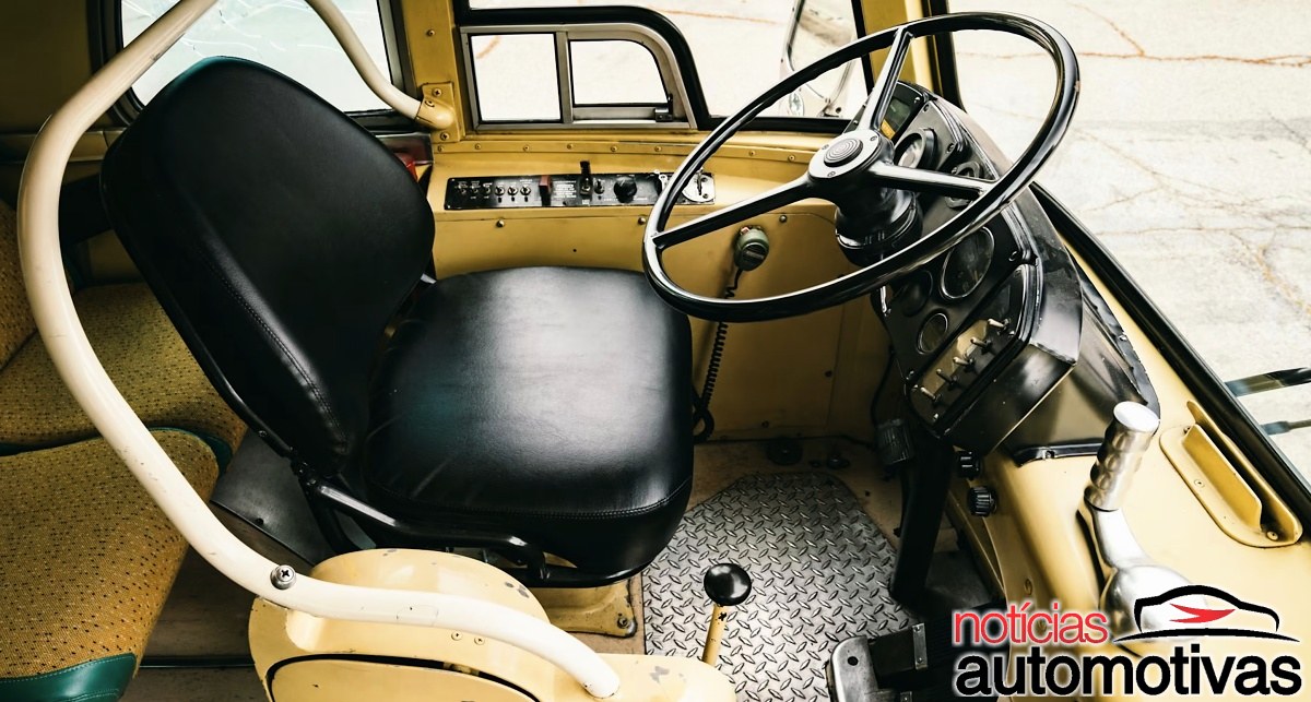 Posto do motorista com banco preto, volante grande, painel e alavanca de câmbio no interior do ônibus.