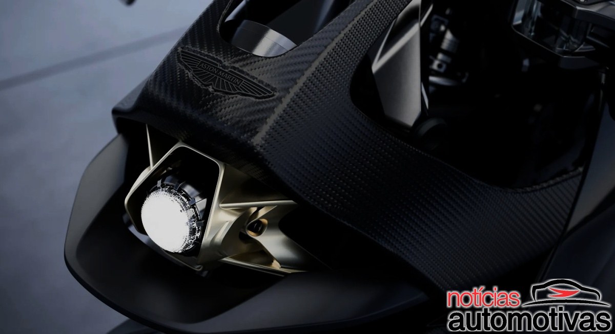 Detalhe frontal de motocicleta preta com peça de fibra de carbono, emblema Aston Martin e farol aceso.