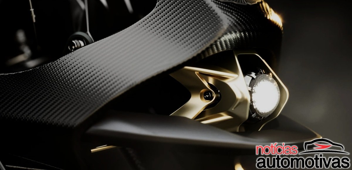 Detalhe de uma motocicleta preta com superfície de fibra de carbono e farol aceso em estrutura dourada.