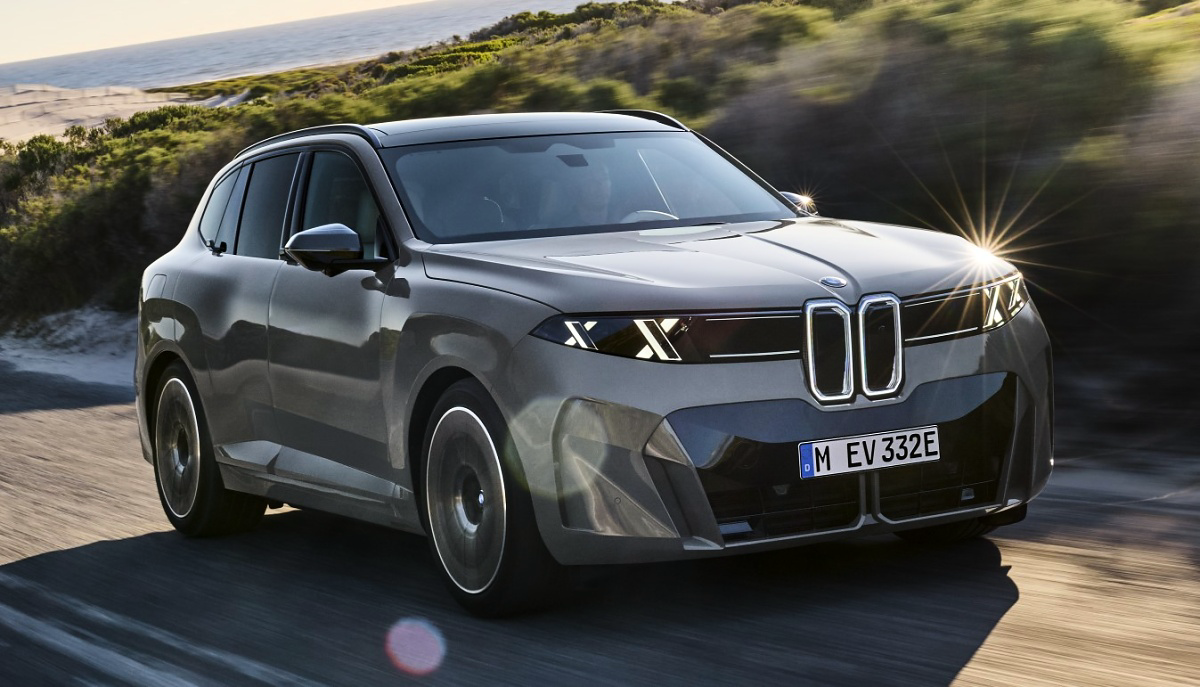 BMW iX5 cinza em uma estrada litorânea, visto de frente e de lado, com vegetação e mar ao fundo.