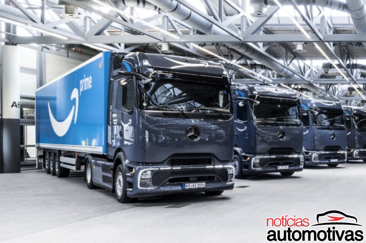 Caminhões Mercedes-Benz azul-escuros com baús azuis da Amazon alinhados dentro de um galpão.