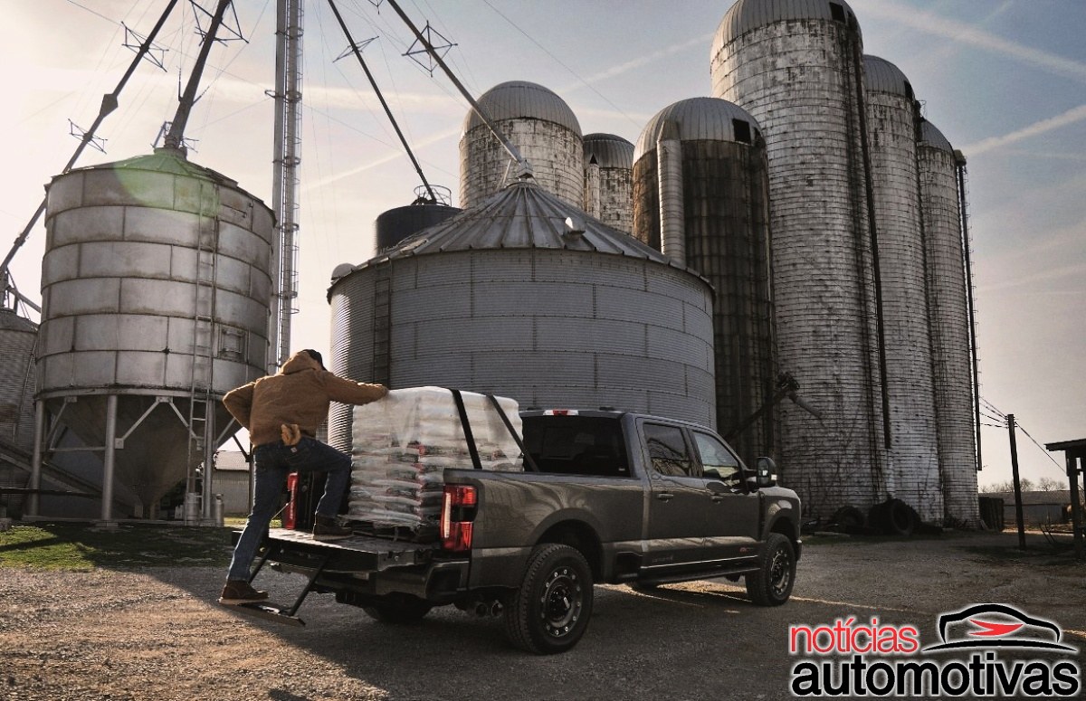 Pessoa coloca um pacote na caçamba de uma picape cinza diante de silos metálicos.