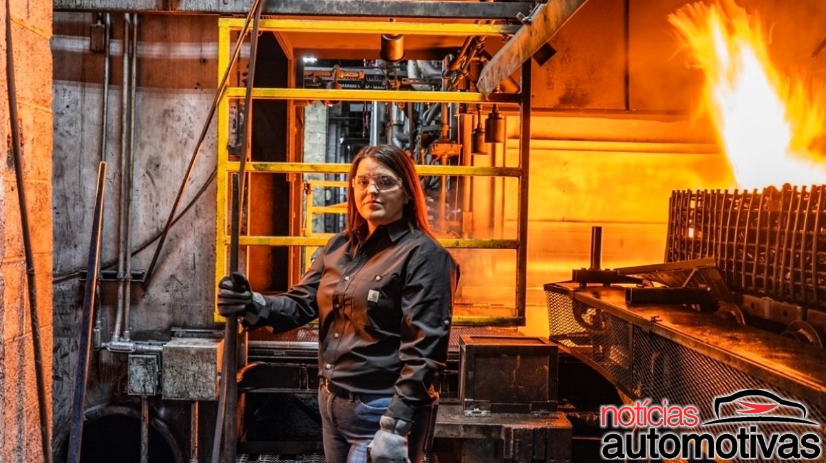 Mulher de óculos e roupas escuras segura uma haste diante de equipamentos industriais e uma chama.