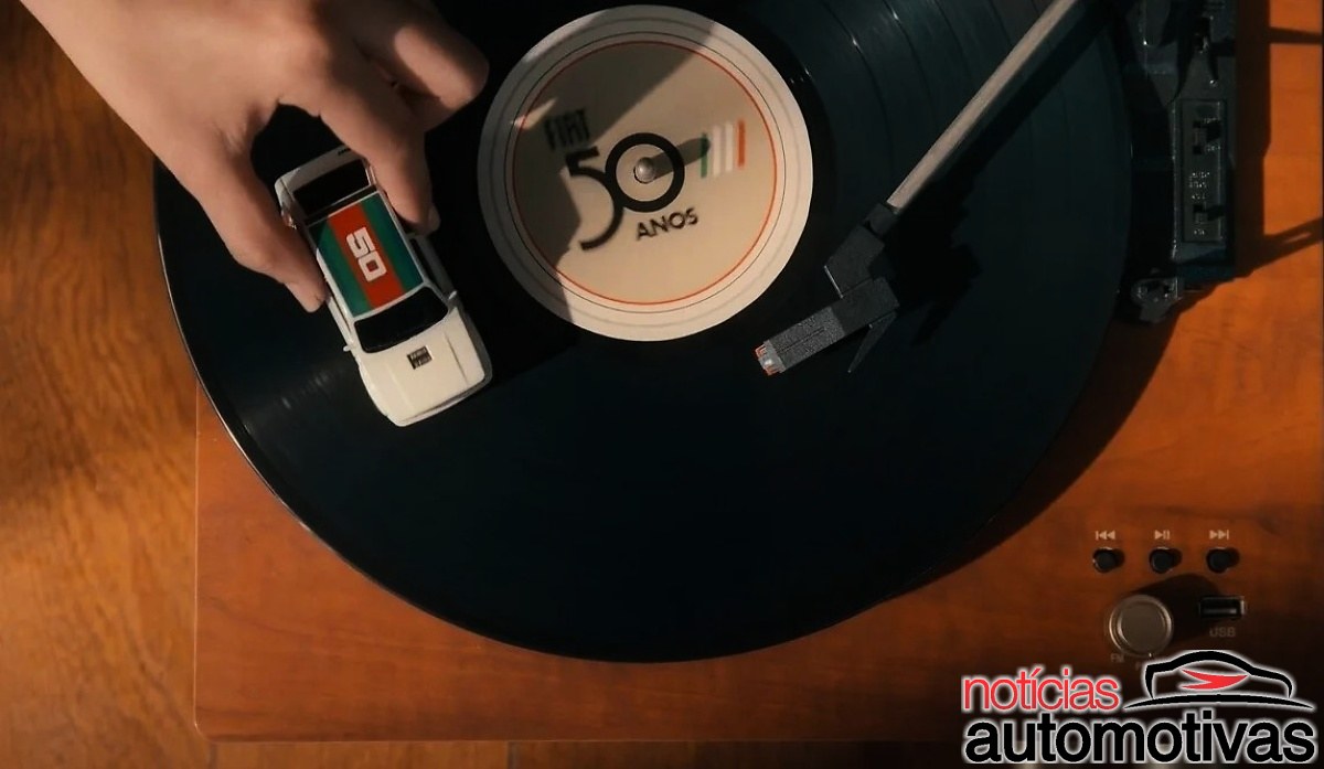 Mão segurando uma miniatura branca de picape sobre um toca-discos, ao lado de um disco com a inscrição Fiat 50 anos.