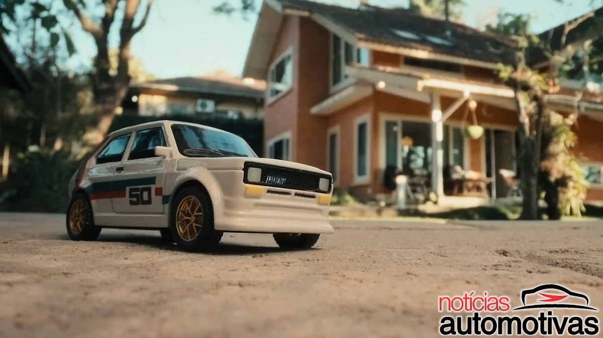 Miniatura branca de um hatch Fiat com adesivo 50 nas laterais, estacionada diante de uma casa.