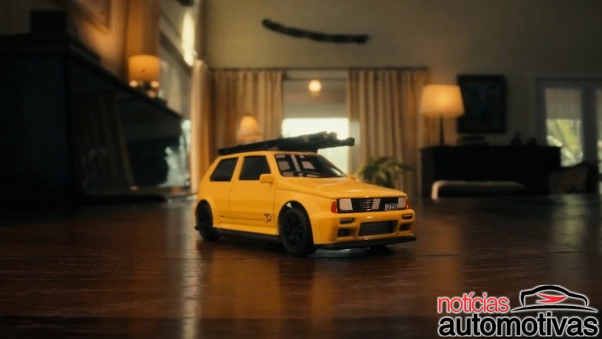 Miniatura amarela de um hatch Fiat com uma escada preta no teto, sobre piso de madeira em uma sala.