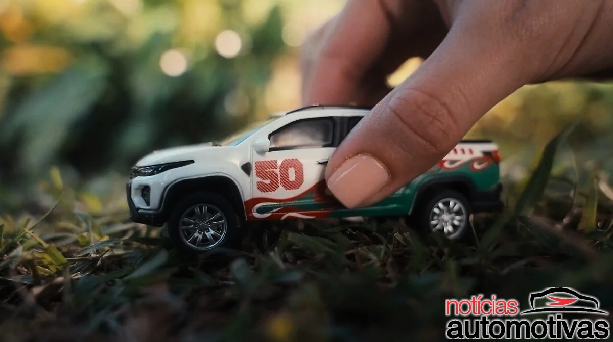 Mão segurando uma miniatura branca de picape Fiat com faixas verdes, vermelhas e o número 50 sobre a grama.