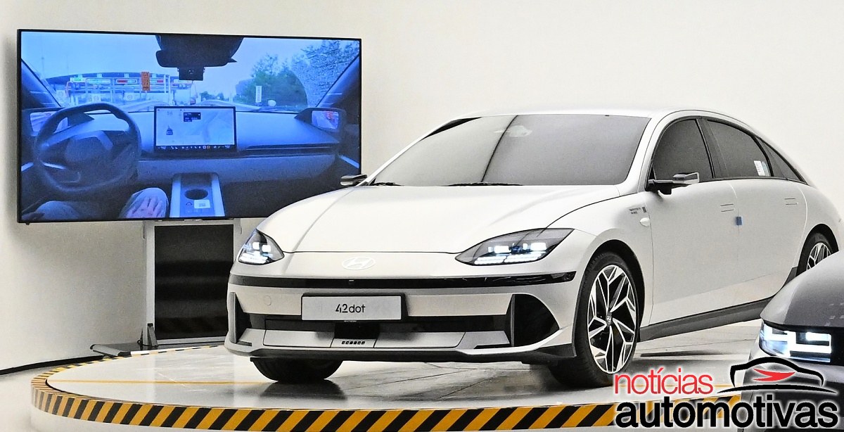 Sedã Hyundai branco em uma plataforma, diante de uma tela que mostra o interior do veículo e uma via urbana.