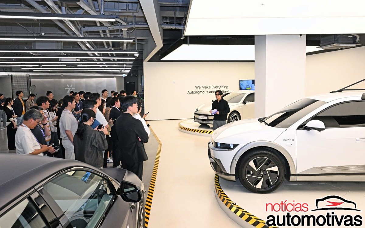 Pessoas observam veículos Hyundai em uma sala de exposição, onde há carros brancos e escuros em plataformas sinalizadas.