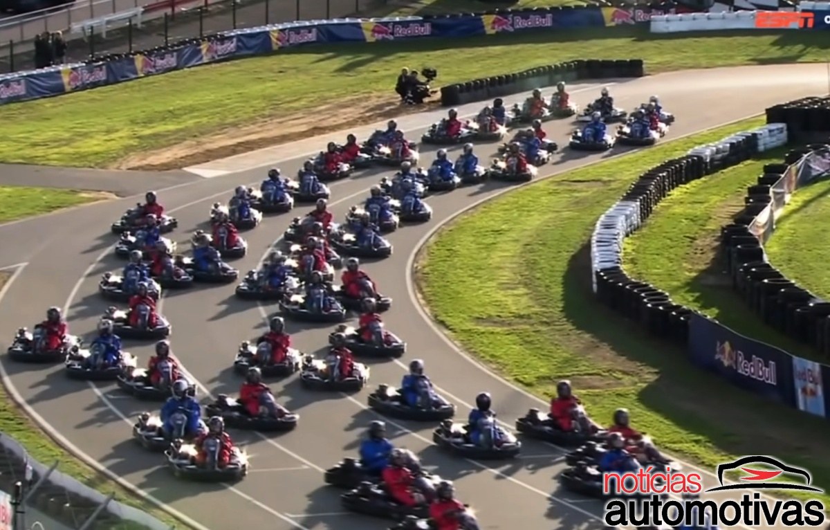 Dezenas de karts com pilotos de macacões azuis e vermelhos contornam uma curva cercada por gramado e barreiras de pneus.