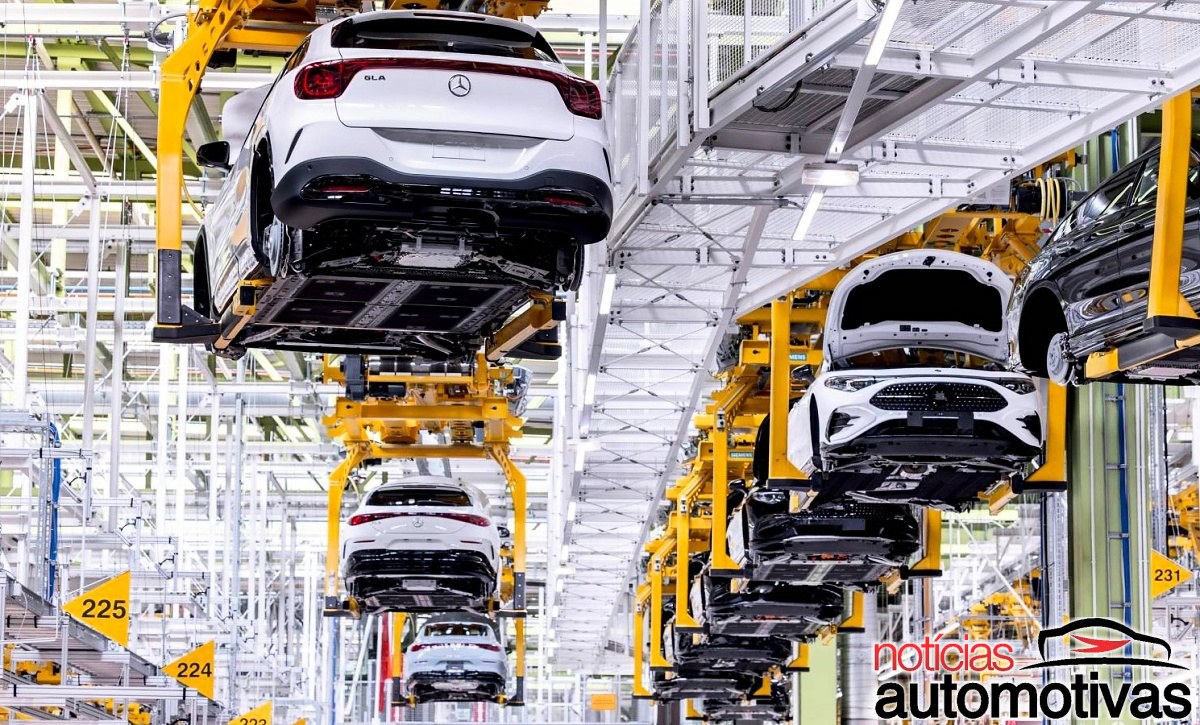 Linha de montagem com vários veículos suspensos em estruturas amarelas dentro de uma fábrica.