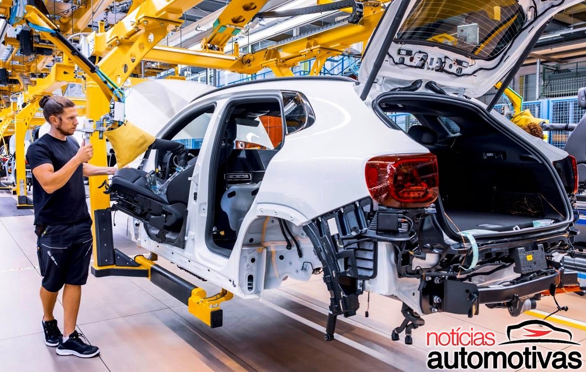 Funcionário trabalha ao lado de uma carroceria branca de SUV sem portas e acabamento, em uma linha de montagem industrial.