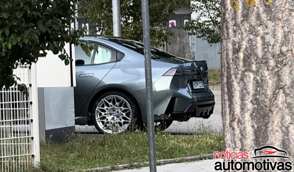 Sedã BMW cinza-escuro parcialmente encoberto por árvores, visto de trás e de lado, com rodas prateadas e quatro saídas de escape.