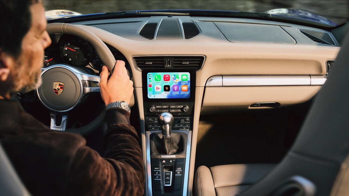 Interior de um Porsche 911 com painel bege, câmbio manual, tela multimídia com ícones do Apple CarPlay e uma pessoa ao volante.