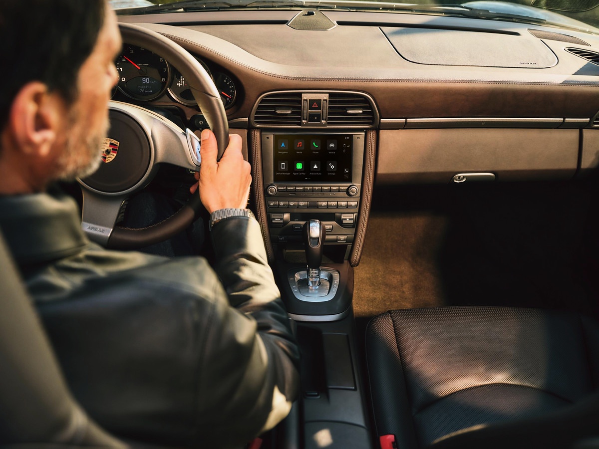 Interior de um Porsche 911 com painel marrom, câmbio automático, tela multimídia e uma pessoa ao volante.
