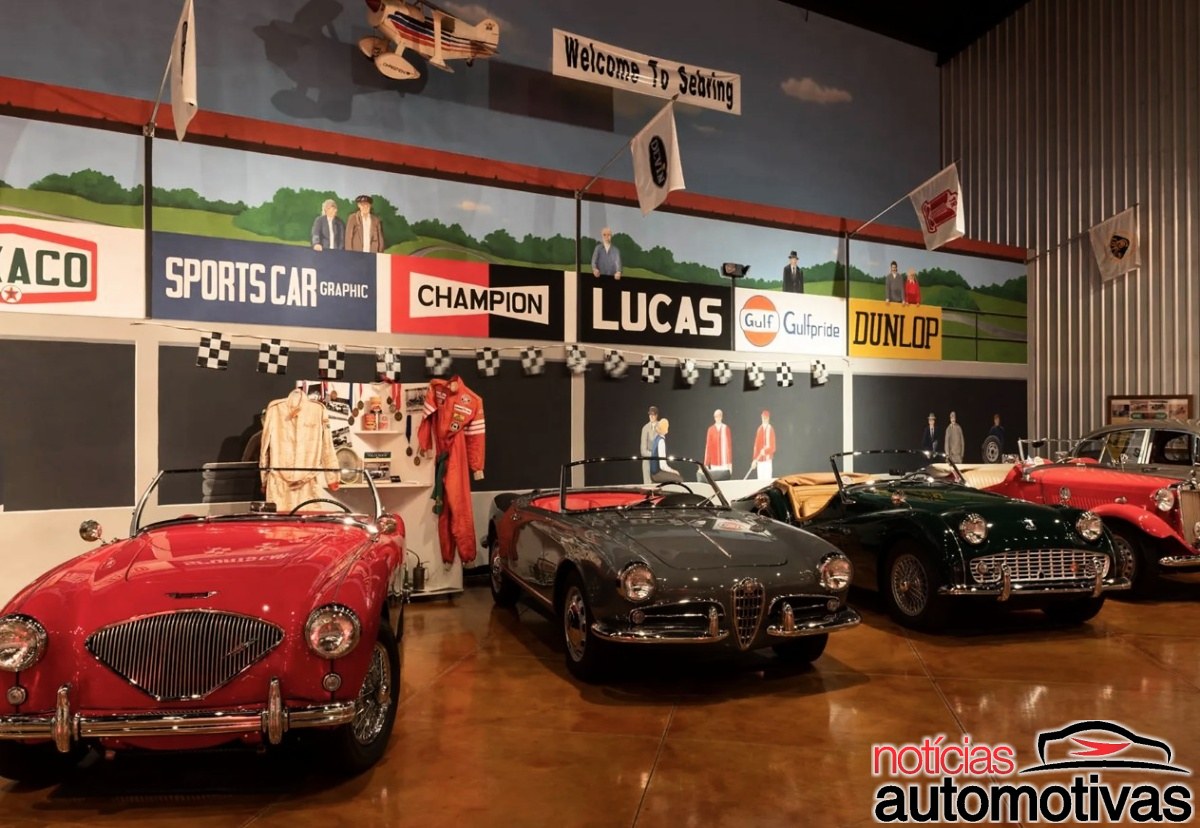 Interior com quatro carros esportivos clássicos, bandeiras, macacões e placas de marcas automotivas nas paredes.