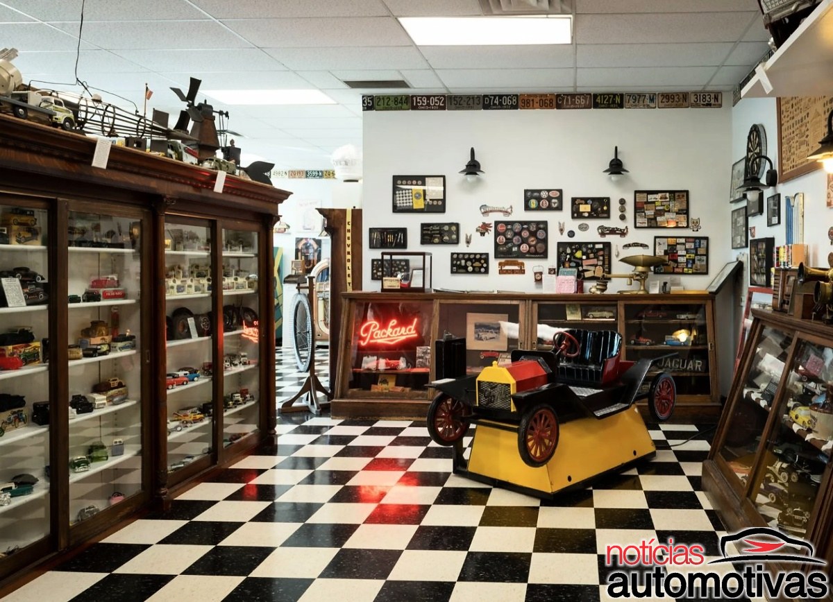 Sala com vitrines de miniaturas, placas decorativas, objetos automobilísticos e um pequeno carro sobre uma base amarela.