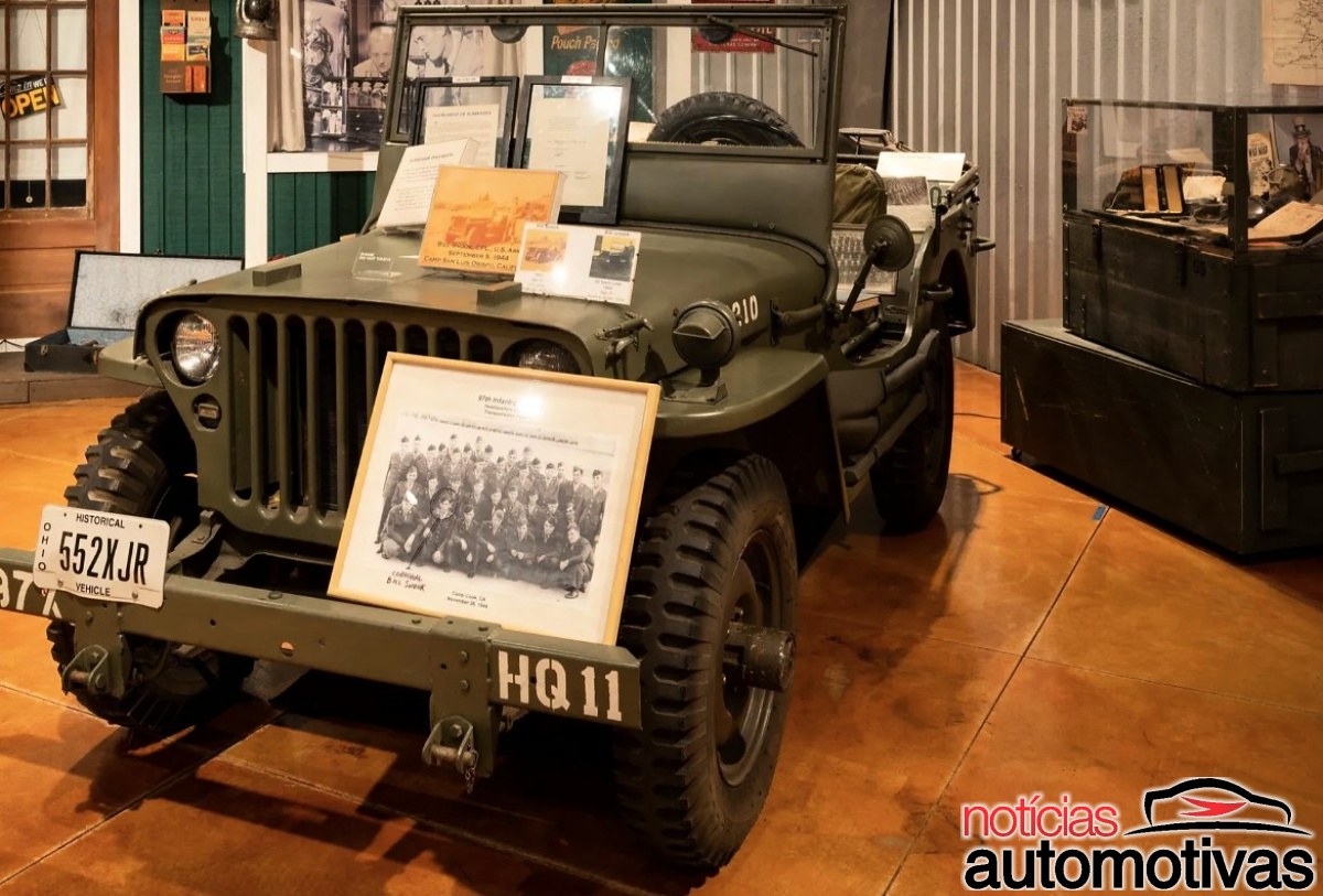 Jipe militar verde-oliva em exposição, com placa histórica, documentos e uma fotografia apoiados à frente.