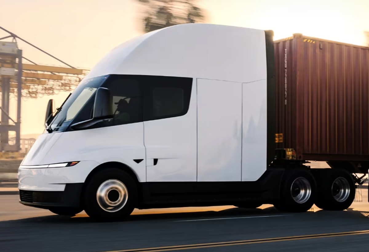 Caminhão Tesla Semi branco visto de lado, rebocando um contêiner marrom em uma área industrial.