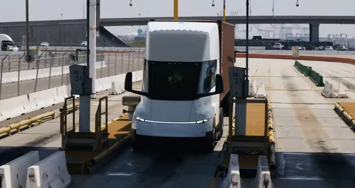 Caminhão Tesla Semi branco com contêiner marrom trafegando entre barreiras e equipamentos de uma pista.