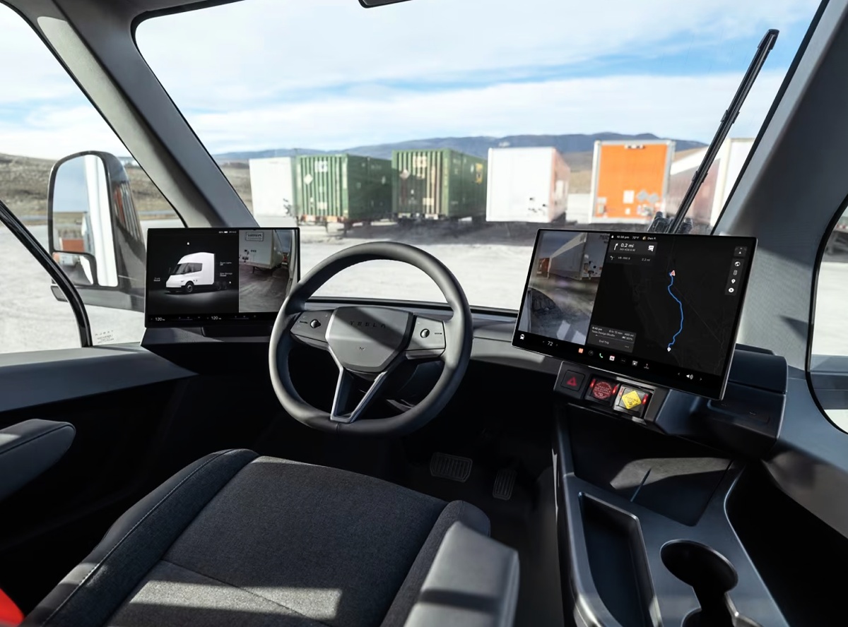 Interior da cabine de um caminhão Tesla Semi, com volante, duas telas digitais e contêineres visíveis à frente.