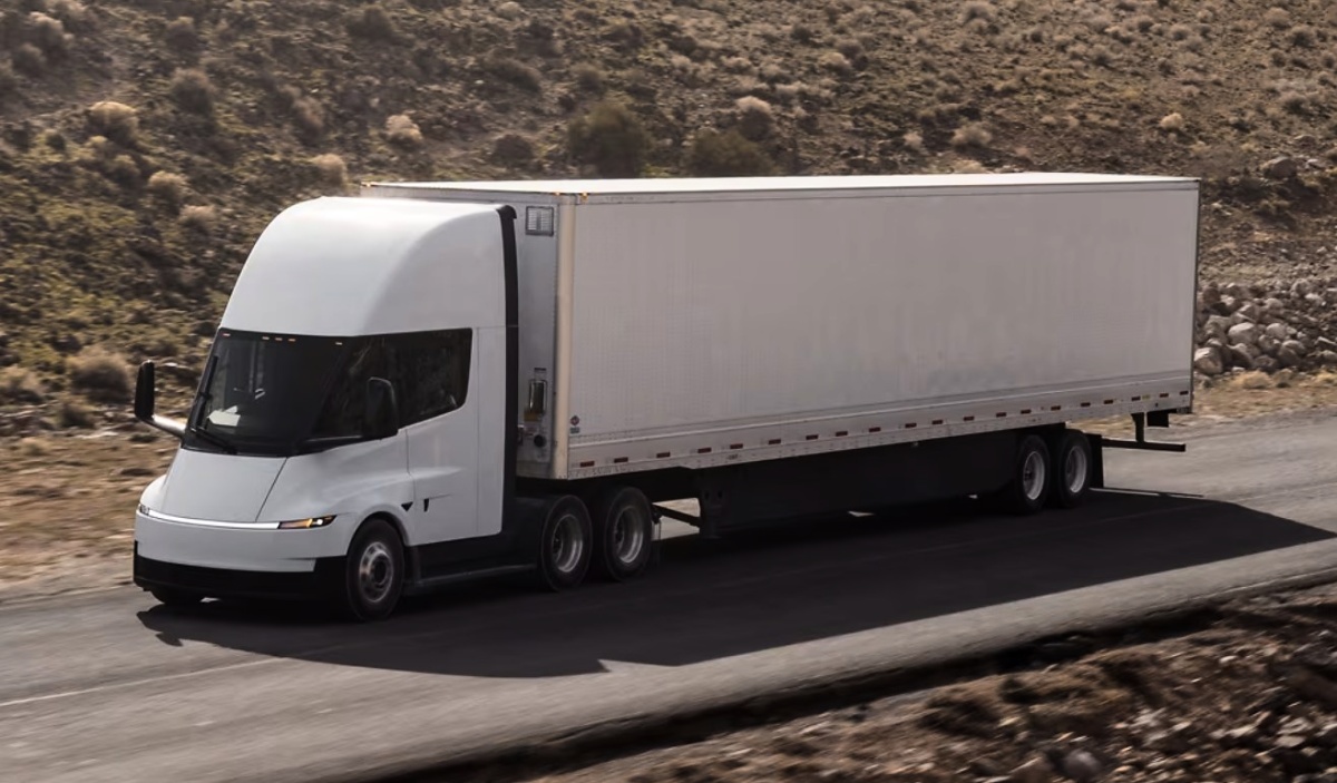 Caminhão Tesla Semi branco com semirreboque fechado trafegando por uma estrada cercada por encostas.