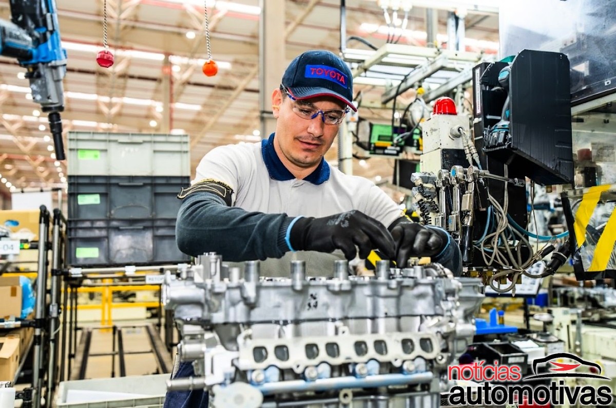 Trabalhador usando óculos de proteção, luvas e boné da Toyota durante a montagem de um motor em uma linha industrial.