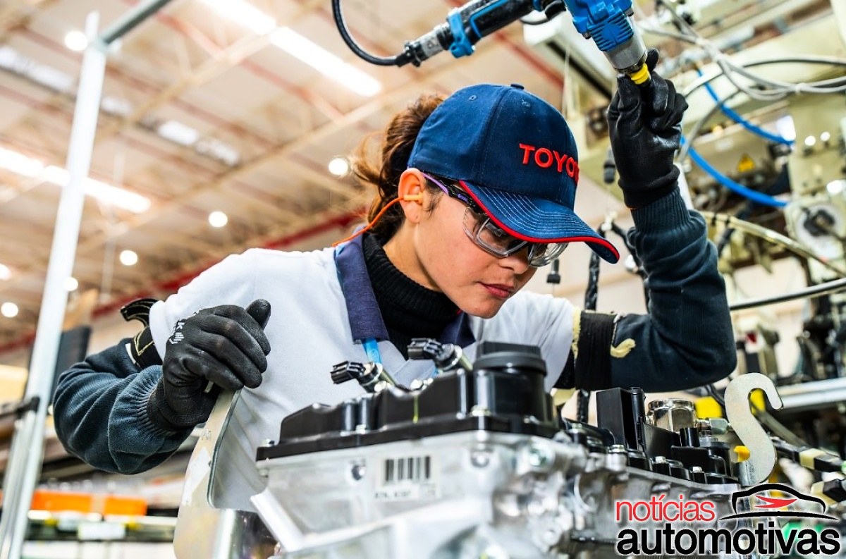 Trabalhadora usando óculos de proteção, luvas e boné da Toyota durante a montagem de um componente em uma linha industrial.