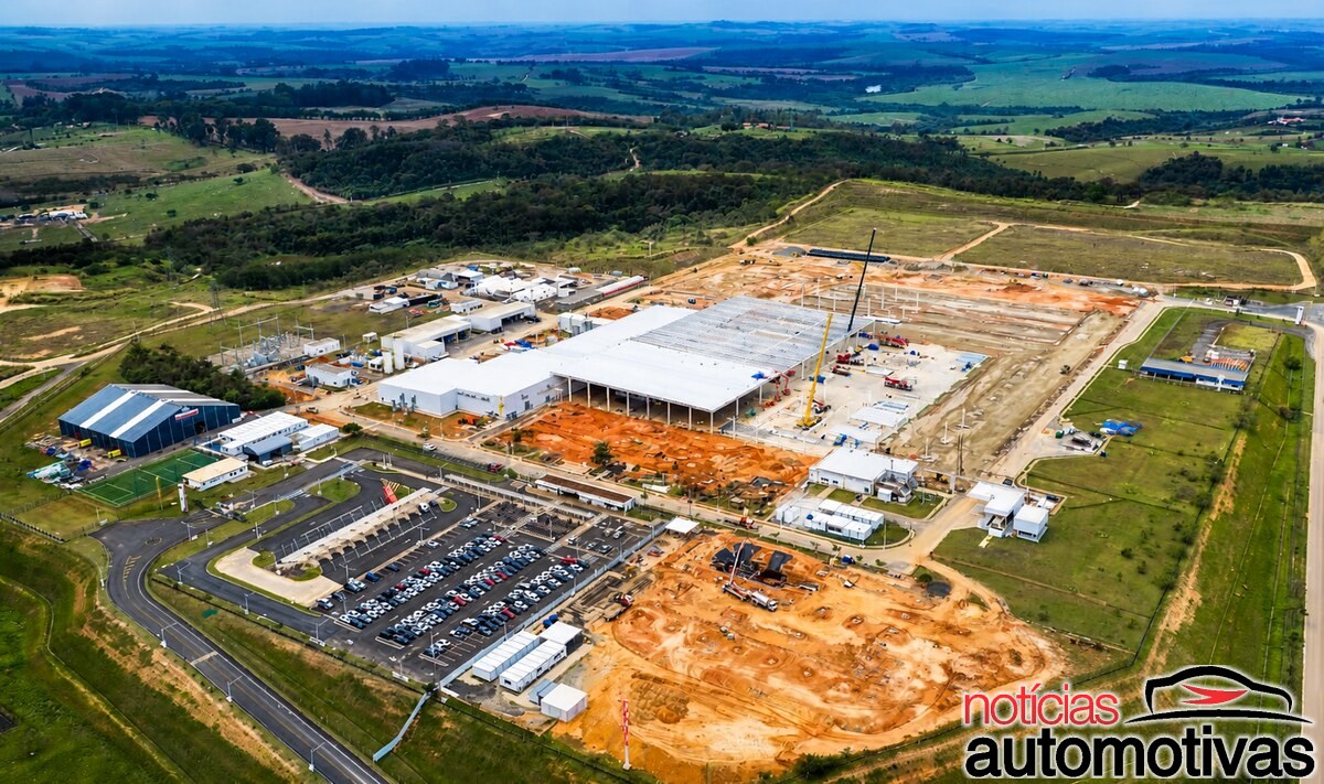 Vista aérea de um complexo industrial em construção, com galpões, áreas de terraplenagem, estacionamento e campos ao redor.