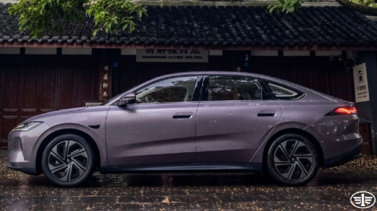 Sedã roxo visto de lado, estacionado diante de uma construção tradicional com árvores ao fundo.