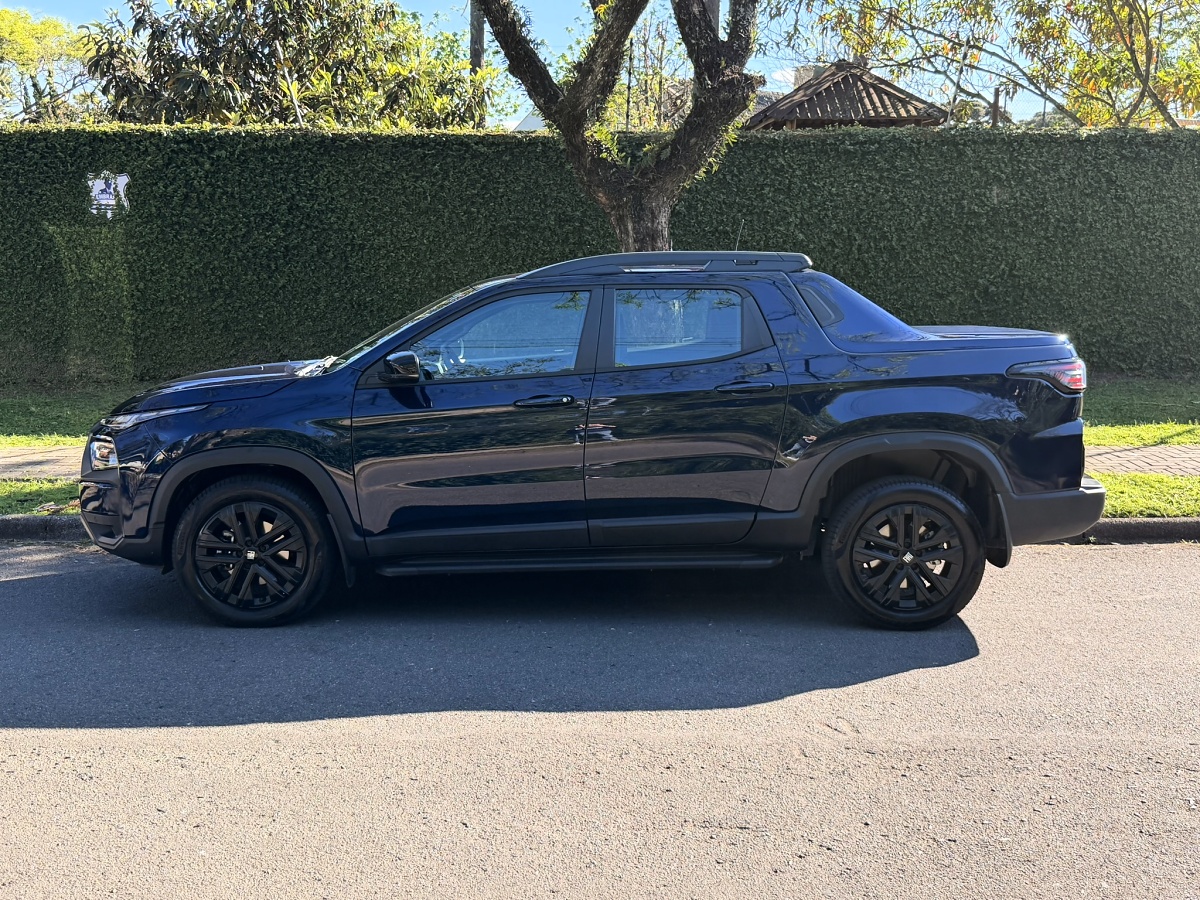 Fiat Toro azul-escura vista de lado, com rodas pretas, estacionada diante de uma cerca viva.