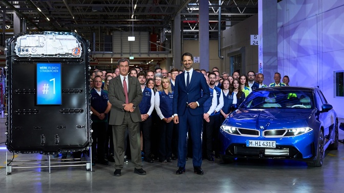 Funcionários da BMW posam ao lado de uma bateria preta e de um BMW azul dentro da fábrica.
