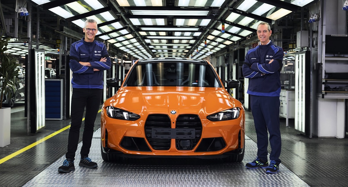 Dois homens em uniforme azul estão ao lado de um BMW M3 laranja dentro de uma fábrica.