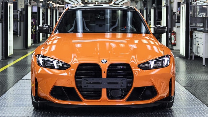 BMW M3 laranja visto de frente no interior de uma fábrica, com máquinas e equipamentos ao fundo.