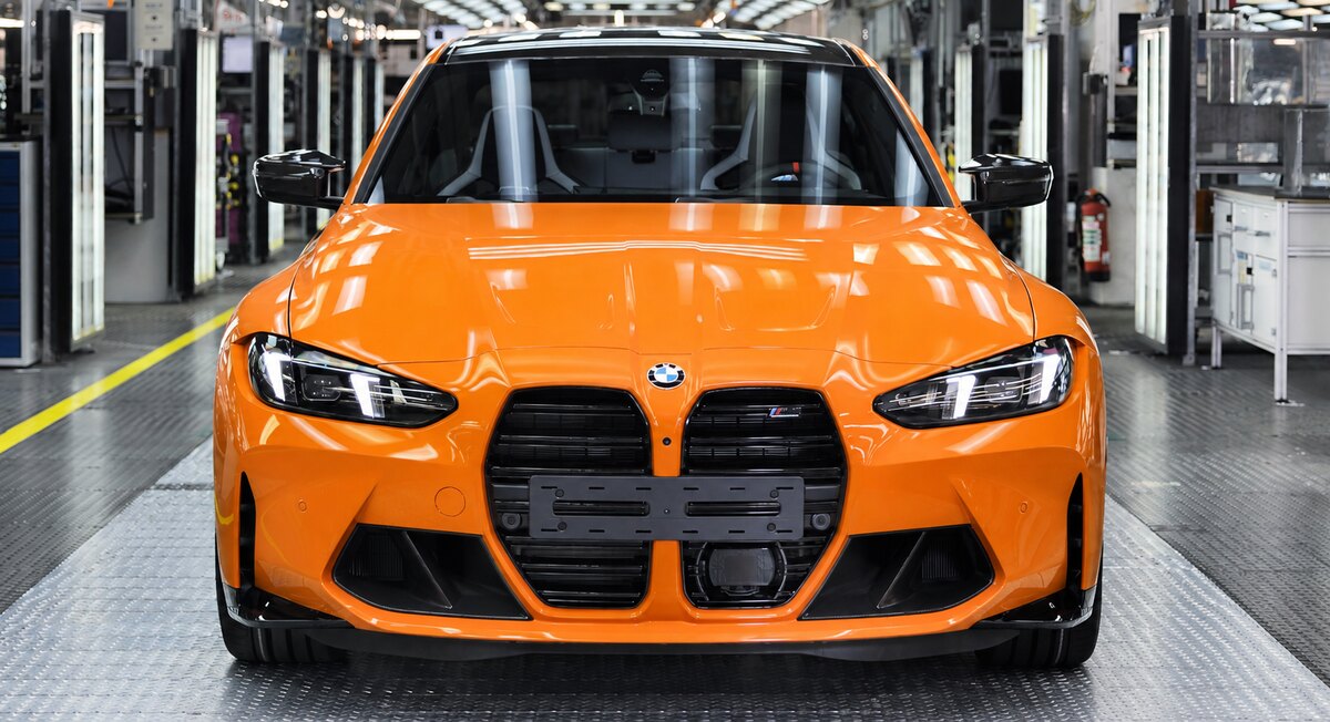 BMW M3 laranja visto de frente no interior de uma fábrica, com máquinas e equipamentos ao fundo.