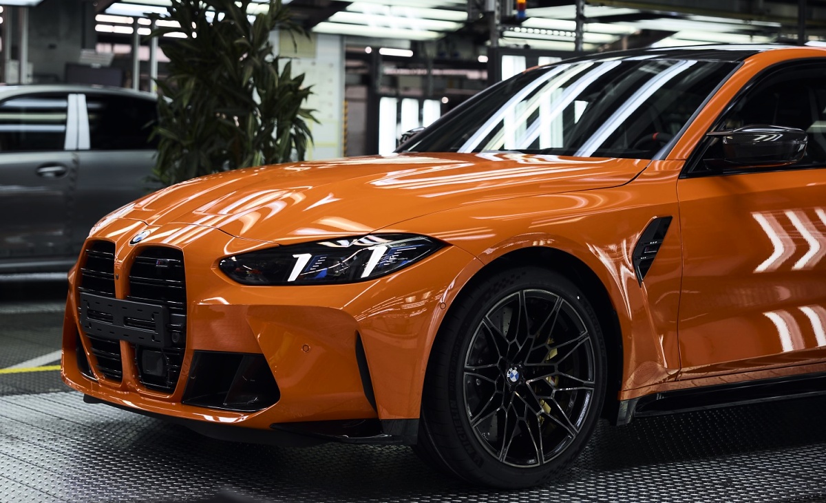 Parte dianteira e lateral de um BMW M3 laranja, com roda preta, dentro de uma fábrica.