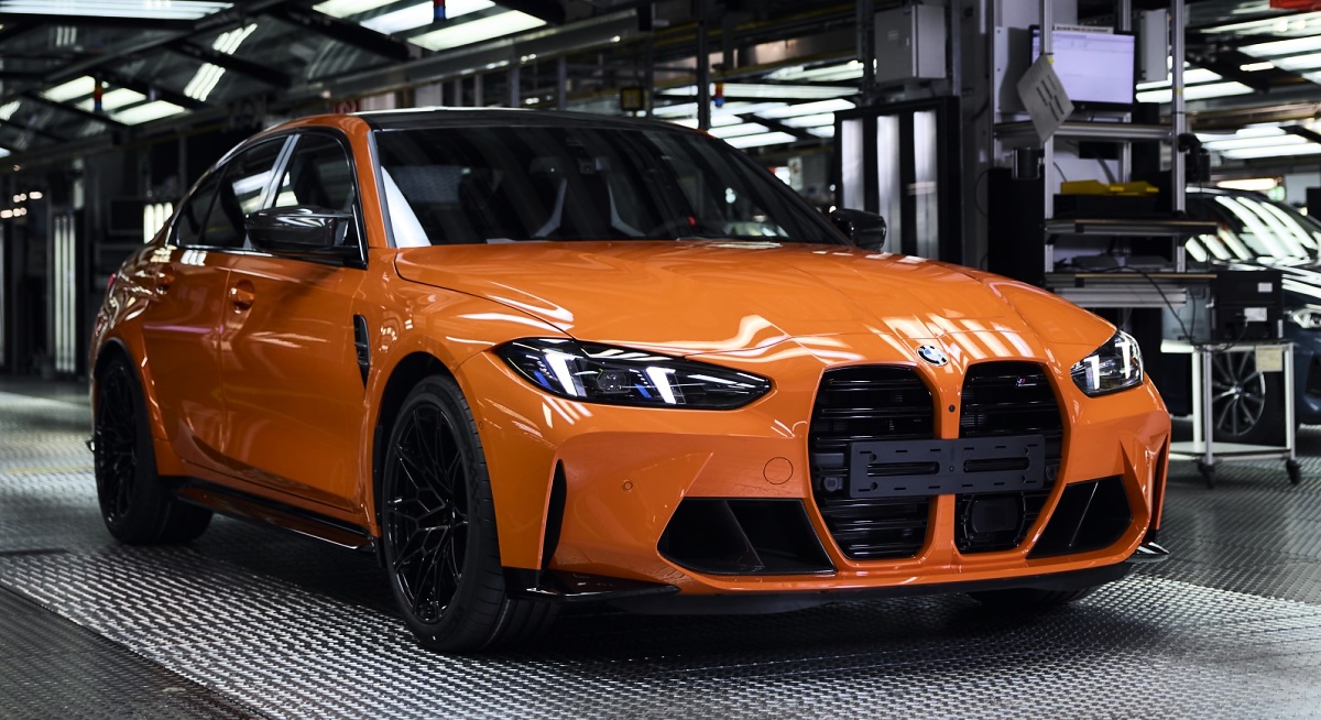 BMW M3 laranja visto de frente e de lado dentro de uma fábrica, com rodas pretas.