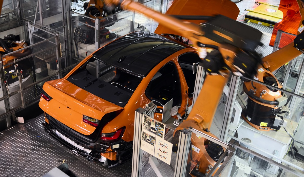 Carroceria laranja de um BMW em uma linha de montagem, cercada por braços robóticos e equipamentos industriais.