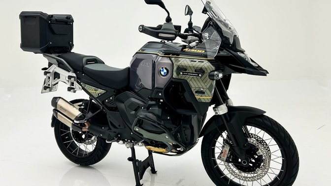 BMW R 1300 GS Adventure preta e cinza, com detalhes amarelos, para-brisa alto e baú traseiro, em fundo branco.
