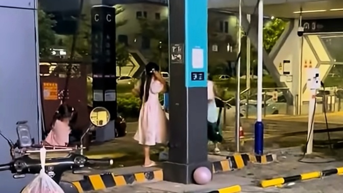 À noite, duas crianças estão junto a um carregador azul e preto; uma usa vestido rosa e a outra está sentada em uma scooter.
