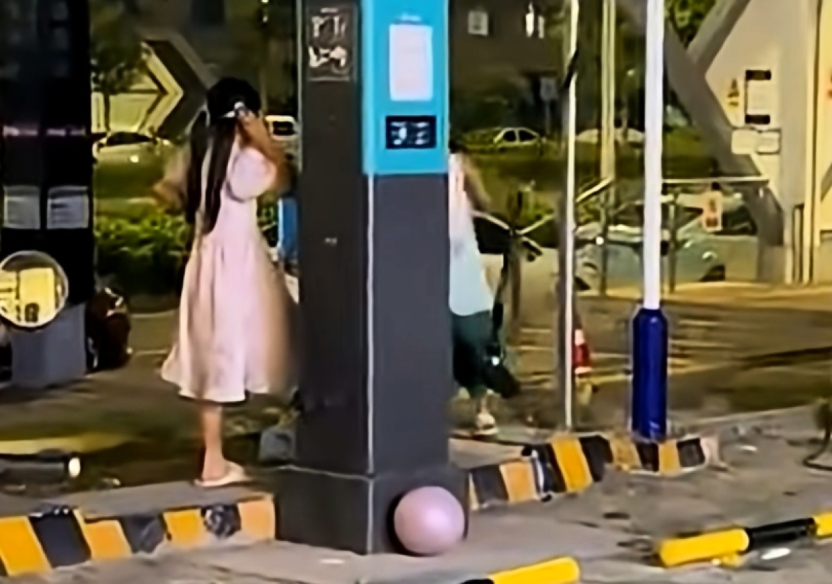 Menina de vestido rosa ao lado de uma coluna azul e preta de recarga; outra pessoa aparece parcialmente atrás do equipamento.