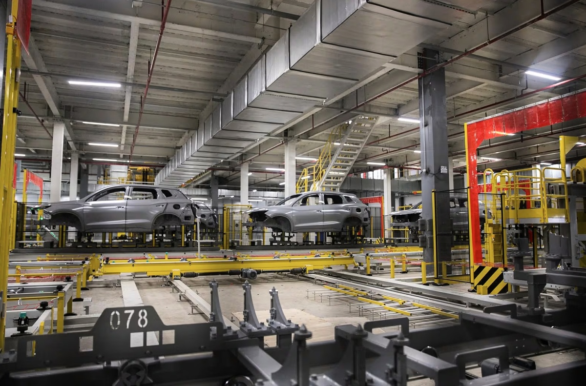 Carrocerias metálicas sem pintura em uma linha industrial com estruturas e equipamentos amarelos.