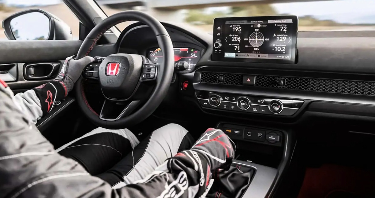 Interior de um Honda Civic Type R com volante de emblema vermelho, tela multimídia, câmbio manual e motorista usando luvas.