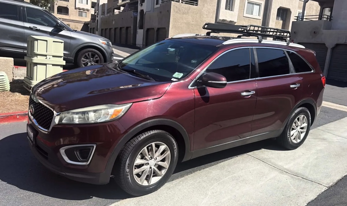 Kia Sorento bordô visto de frente e de lado, com bagageiro de teto, estacionado diante de prédios.