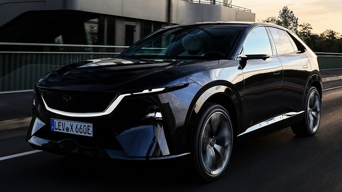 Carro preto da Mazda em movimento sobre uma via, com edifício e guarda-corpo ao fundo.