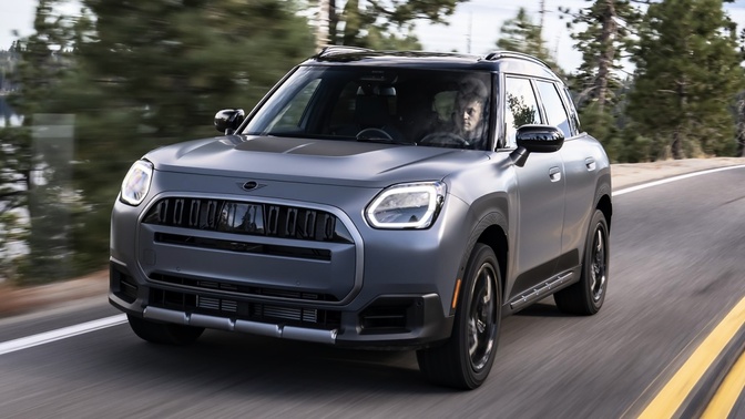 SUV MINI Countryman cinza em uma estrada, com um ocupante ao volante e árvores ao fundo.